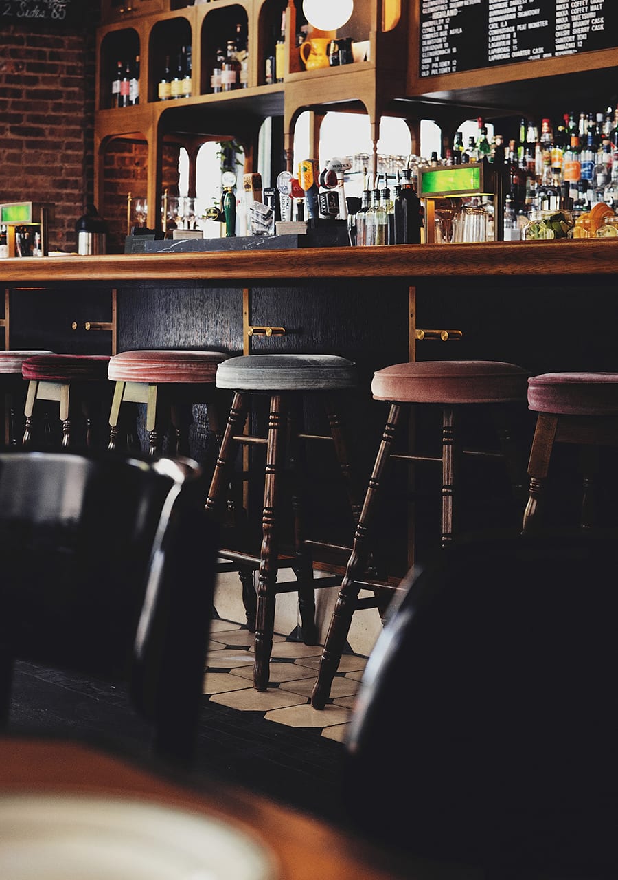 Eine gemütliche Bar mit hohen Holzhockern, einem dunklen Tresen, Flaschen und Gläsern, die fein säuberlich in Regalen angeordnet sind, und einer darüber hängenden Speisekarte. Die Szene ist warm beleuchtet und einladend. Bild der Website bautec-zeitarbeit.de