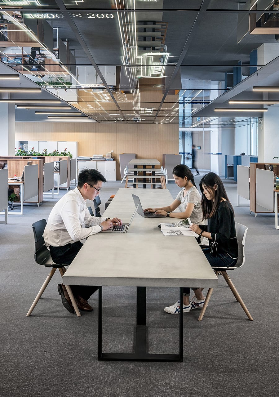 Drei Personen sitzen an einem langen Tisch in einem modernen Großraumbüro, arbeiten an Laptops und lesen Papiere. Der Raum ist hell, mit Glaswänden, eleganten Möbeln und Pflanzen im Hintergrund. Bild der Website bautec-zeitarbeit.de